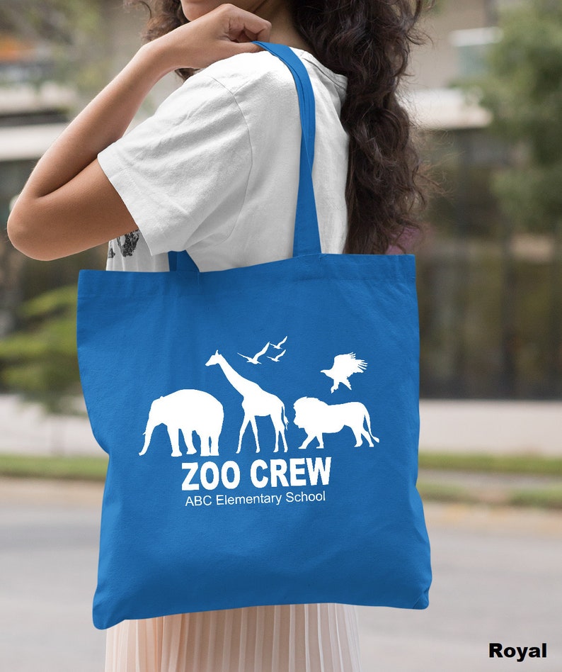 May include: A royal blue tote bag with white silhouettes of animals, including an elephant, giraffe, lion, and birds. The text "Zoo Crew" and "ABC Elementary School" is printed in white on the bag.