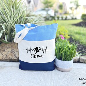 Puede incluir: Una bolsa de mano azul, blanca y azul marino con un fondo blanco y un gr&aacute;fico negro de una aguja de coser y un hilo con una l&iacute;nea de latido del coraz&oacute;n y el nombre "Olivia" impreso en ella.