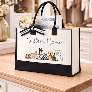 May include: A rectangular canvas tote bag with black trim and handles, featuring a black and white striped ribbon tied at the base of one handle. The front displays the text Custom Name above a colorful illustration of nine various dog breeds.