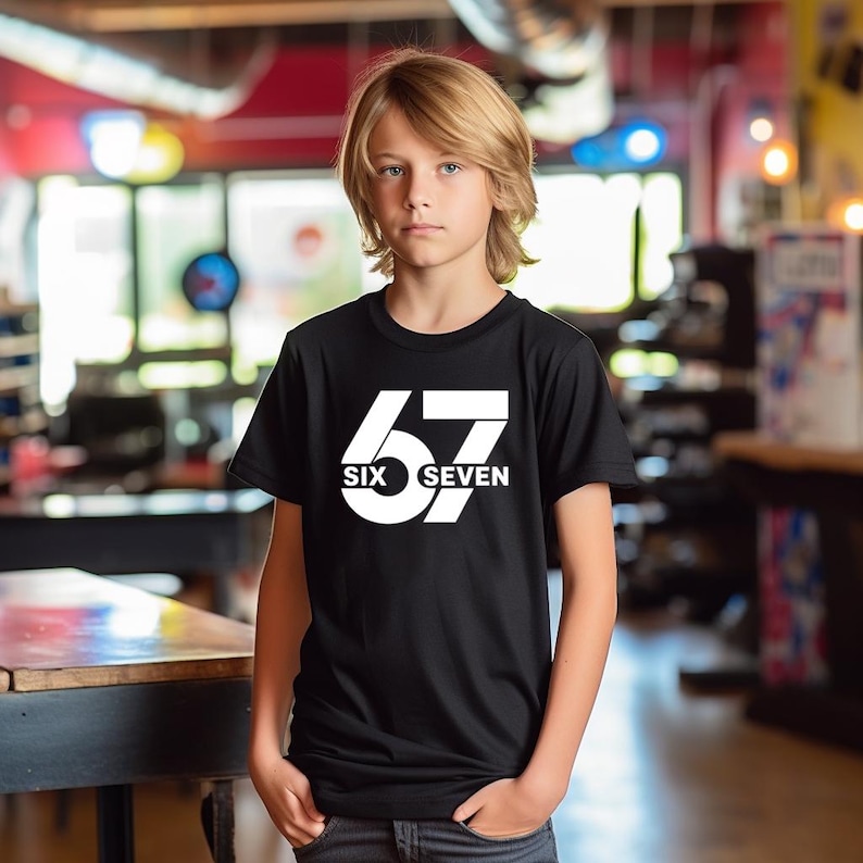 K&ouml;nnte beinhalten: Ein schwarzes T-Shirt mit der wei&szlig;en Zahl "67" und den W&ouml;rtern "SIX" und "SEVEN" darunter. Das T-Shirt wird von einer Person mit blonden Haaren getragen.