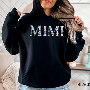 May include: Black hooded sweatshirt with the word "MIMI" in floral print across the chest. The letters are filled with a pattern of small blue, pink, and white flowers. The word "BLACK" is printed in white at the bottom right.