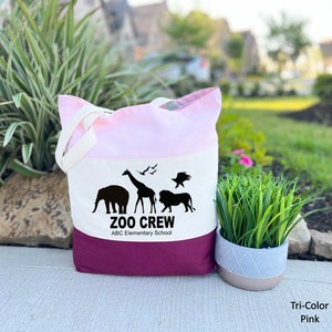 May include: A tri-color tote bag with a pink, white, and burgundy design. The bag features a black silhouette of a giraffe, elephant, lion, and birds with the text "Zoo Crew ABC Elementary School".