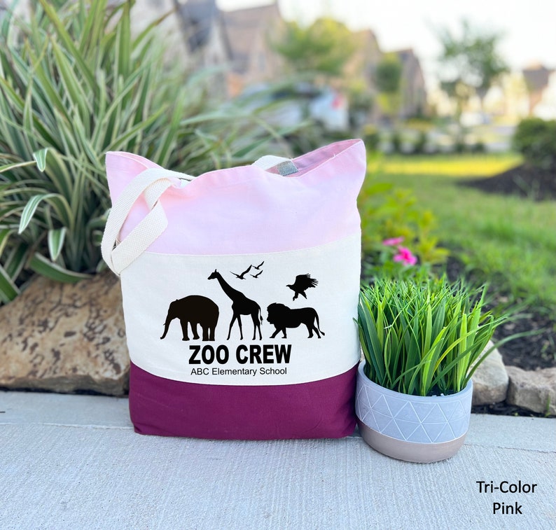 May include: A tri-color tote bag with a pink, white, and burgundy design. The bag features a black silhouette of a giraffe, elephant, lion, and birds with the text "Zoo Crew ABC Elementary School".