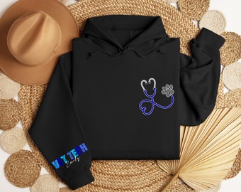 May include: Black hooded sweatshirt with a blue stethoscope and paw print design. The text "VET TECH Emily" is on the left sleeve.