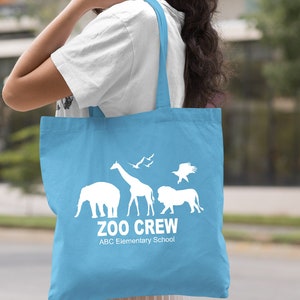 May include: Light blue tote bag with white silhouette of animals, including an elephant, giraffe, lion, and birds. The text "Zoo Crew" and "ABC Elementary School" is printed on the bag.