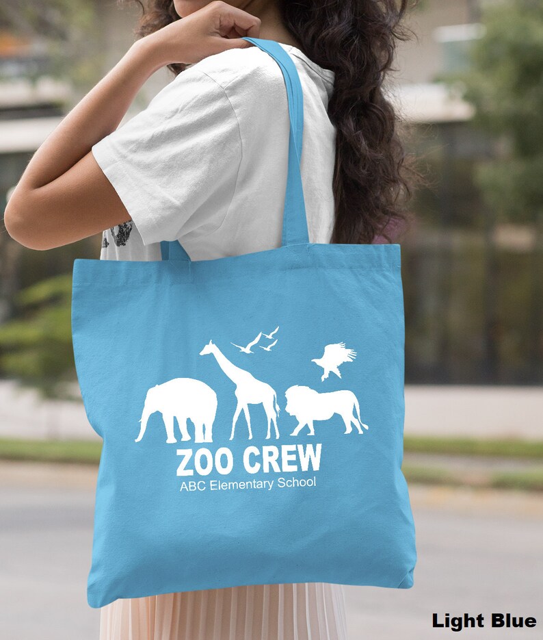May include: Light blue tote bag with white silhouette of animals, including an elephant, giraffe, lion, and birds. The text "Zoo Crew" and "ABC Elementary School" is printed on the bag.