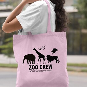 May include: Light pink tote bag with black silhouettes of animals, including an elephant, giraffe, lion, and eagle. The text "Zoo Crew" and "ABC Elementary School" is printed on the bag.