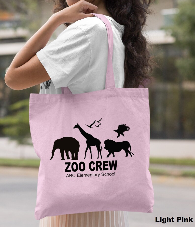May include: Light pink tote bag with black silhouettes of animals, including an elephant, giraffe, lion, and eagle. The text "Zoo Crew" and "ABC Elementary School" is printed on the bag.