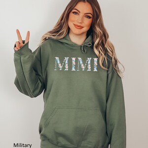 May include: Military green hoodie with the word "MIMI" in a floral design across the chest. The hoodie has a drawstring hood and a front pocket. The text "Military Green" is at the bottom left.