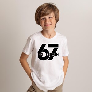 K&ouml;nnte beinhalten: Ein wei&szlig;es T-Shirt mit der Zahl "67" in fetter schwarzer Schrift. Die W&ouml;rter "SIX" und "SEVEN" befinden sich unter den Ziffern. Das T-Shirt wird von einer jungen Person getragen.