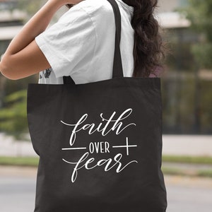 Może przedstawiać: Czarna torba typu tote z białym napisem "faith over fear" napisanym kursywą.
