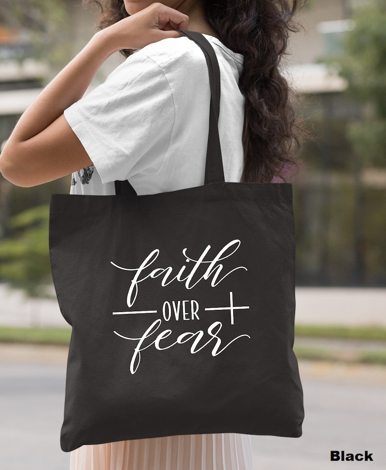 Może przedstawiać: Czarna torba typu tote z białym napisem "faith over fear" napisanym kursywą.