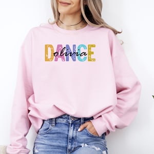 May include: A light pink sweatshirt with the word "DANCE" in colorful block letters and the name "Olivia" in cursive script. The sweatshirt is made of a soft material and is suitable for casual wear.