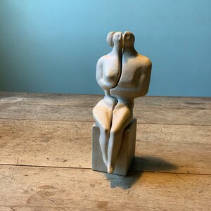 May include: A white ceramic sculpture of two figures, joined together, sitting on a square base. The sculpture is abstract and modern in style.