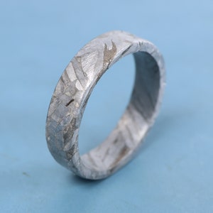 May include: A silver ring crafted from meteorite material. The ring features a distinctive, textured surface with a pattern of lines and swirls.