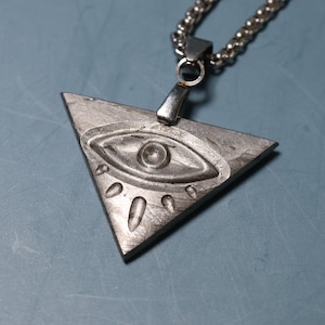Peut inclure: Un collier avec un pendentif en forme de triangle argenté orné d'un motif d'œil. L'œil est détaillé avec une pupille en relief et des cils.