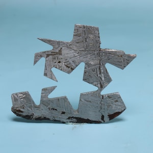 May include: A cut, irregular-shaped piece of metal with a star-shaped cutout. The metal has a silver and grey colour with a visible crystalline pattern and a crack running through the centre. The object is set against a light blue background.