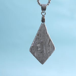 Puede incluir: Un collar con un colgante de plata en forma de diamante, hecho de meteorito con una superficie texturizada única.