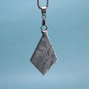 Puede incluir: Un colgante en forma de diamante con un patrón texturizado único, suspendido de una cadena plateada. El colgante es de color gris metálico y probablemente está hecho de piedra o metal. El fondo es azul claro liso.