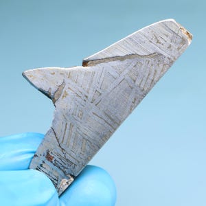 Puede incluir: Primer plano de un meteorito de color plateado con una superficie rugosa y texturizada. El meteorito tiene un patrón de Widmanstätten distintivo, que es una característica de los meteoritos de hierro.