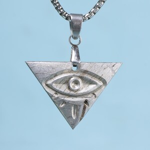Puede incluir: Un collar con un colgante de triángulo plateado con un diseño de ojo. El colgante está hecho de metal y tiene una superficie texturizada.