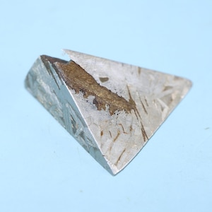 May include: A triangular-shaped, metallic object with a weathered appearance. The object has a silver-gray color with brown and dark streaks. The edges are sharp, and the surface shows a unique pattern. The object is set against a light blue background.