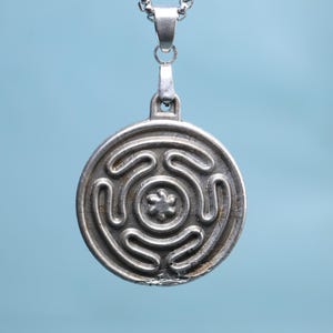 Può includere: Collana con pendente argentato con un design circolare con un motivo a labirinto e un elemento floreale centrale. Il pendente è appeso a una delicata catena su uno sfondo azzurro. Il gioiello è un accessorio.