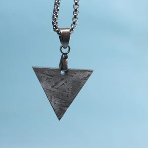 Puede incluir: Collar plateado con un colgante triangular. El colgante, con superficie texturizada, cuelga de una cadena. El fondo es de color azul claro.