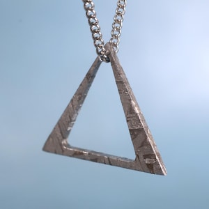 Puede incluir: Un collar con un colgante triangular de plata con superficie texturizada. El colgante está hecho de material de meteorito.