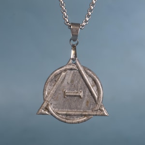 Puede incluir: Un collar de plata con un colgante que presenta un triángulo dentro de un círculo. El colgante tiene una superficie texturizada y una pequeña barra en el centro del triángulo.