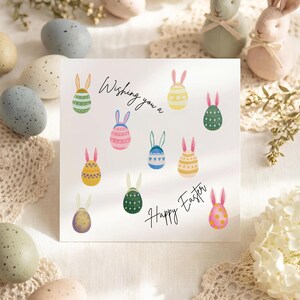 May include: A square Easter card with the text "Wishing you a Happy Easter" and illustrations of colourful Easter eggs with bunny ears. Decorative Easter eggs and bunny figurines surround the card.