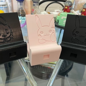 May include: Three 3D printed phone stands, each featuring a different Pokemon character. The stands are black, pink, and black. The Pokemon characters are Eevee, Pikachu, and Charmander.