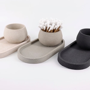 Könnte beinhalten: Drei ovale Betontabletts in Beige, Grau und Schwarz mit passenden runden Schalen. Die mittlere Schale enthält Wattestäbchen.