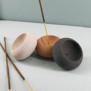 Op de afbeelding: Drie kleine, gespikkelde keramische wierookhouders in beige, bruin en zwart. Wierookstokjes zijn in de houders geplaatst, met twee stokjes op het oppervlak. De houders hebben een afgeronde, komvormige vorm.