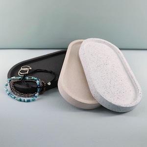 Op de afbeelding: Drie ovale trays in zwart, beige en gespikkeld wit. Een zwarte tray bevat ringen en armbanden met blauwe en groene kralen. De trays zijn waarschijnlijk voor sieraden of kleine items.