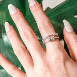 May include: A close-up of a hand wearing a silver ring with multiple bands of small diamonds. The ring is on the middle finger of the hand. The hand is resting on a green leaf.