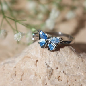 May include: A silver butterfly ring with light blue gemstones. The butterfly design features a detailed wing structure with multiple blue stones. The ring is set on a light-coloured, textured surface, with soft focus greenery in the background.