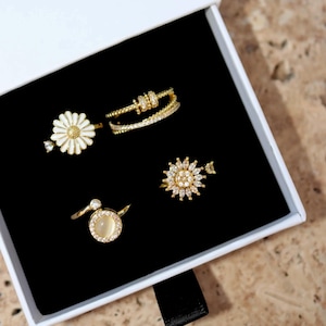 May include: A set of four gold rings with white stones. The rings feature a daisy, a circle with a white stone, and two different styles of sunburst designs.