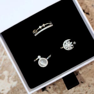 May include: Three silver rings with clear stones. The first ring has a double band with a small bar in the centre. The second ring has a large round stone with a halo of smaller stones. The third ring has a crescent moon shape with a star in the centre.