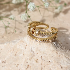 May include: A gold ring with a distinctive design. The ring has multiple bands, one with small, clear stones, and another with a beaded texture. The centre has a rotating element with more clear stones. The ring is set on a light-coloured stone.
