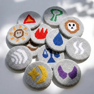 Op de afbeelding: Een verzameling grijze, ronde onderzetters met geborduurde ontwerpen. Elke onderzetter heeft een ander symbool in kleuren als rood, geel, blauw en groen. Ontwerpen zijn onder meer een zon, vuur, een waterdruppel en een maan.