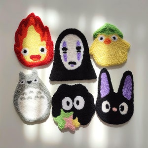 Puede incluir: Seis adornos de alfombra de personajes de fieltro de colores. Los personajes incluyen un pollito amarillo con un sombrero verde, un personaje negro sin rostro de Spirited Away, un Totoro gris, un gato negro con orejas moradas, un espíritu de hollín negro con una estrella rosa y verde, y una llama roja y amarilla.