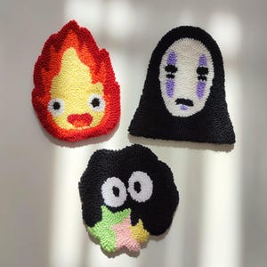 May include: Three handmade felt wall hangings featuring characters from the animated film Spirited Away. The first is a red and yellow flame-shaped character with a smiling face. The second is a black and white character with a large, white, faceless mask. The third is a black character with large white eyes and a green, pink, and yellow star.