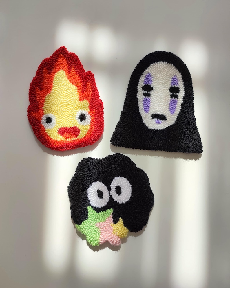 May include: Three handmade felt wall hangings featuring characters from the animated film Spirited Away. The first is a red and yellow flame-shaped character with a smiling face. The second is a black and white character with a large, white, faceless mask. The third is a black character with large white eyes and a green, pink, and yellow star.