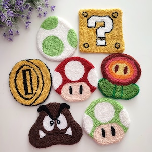 May include: Seven colorful felt coasters featuring iconic characters and items from the Super Mario Bros. video game franchise. The coasters include a green Yoshi egg, a red mushroom, a green mushroom, a brown Goomba, a yellow question block, a gold coin, and a red flower.