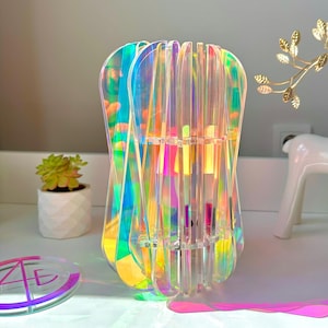 Könnte beinhalten: Eine schillernde Acrylvase mit einem einzigartigen, geschichteten Design. Die Vase reflektiert ein Farbspektrum, darunter Rosa, Gelb und Blau. Eine kleine Sukkulente in einem weißen Topf und eine dekorative Hirschfigur befinden sich im Hintergrund.