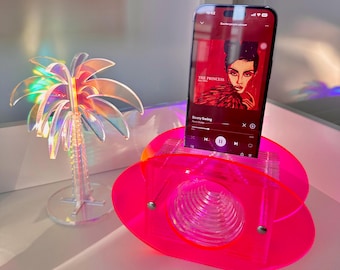 May include: A neon pink acrylic phone stand with a circular design, holding a smartphone displaying a music app. A clear, iridescent palm tree decoration is to the left. The phone screen shows the text "THE PRINCESS" and "Booty Swing".