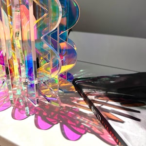 May include: A translucent, iridescent acrylic sculpture with a wavy, multi-layered design that casts vibrant, colorful shadows. Next to it lies a book with a black cover featuring the text "DAVID" and "FIVE HUNDRED" in white lettering.