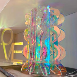 May include: A vertical sculpture constructed from interlocking iridescent acrylic panels. The translucent material displays a spectrum of shifting colors including pink, blue, and yellow, creating a multi-layered, wavy, and geometric structure.