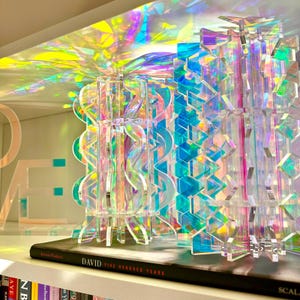 May include: Two iridescent acrylic bookends with geometric and wavy cutouts that reflect vibrant rainbow colors. The left bookend features a repeating wave pattern, while the right displays a jagged, angular design, both resting on a book titled DAVID.